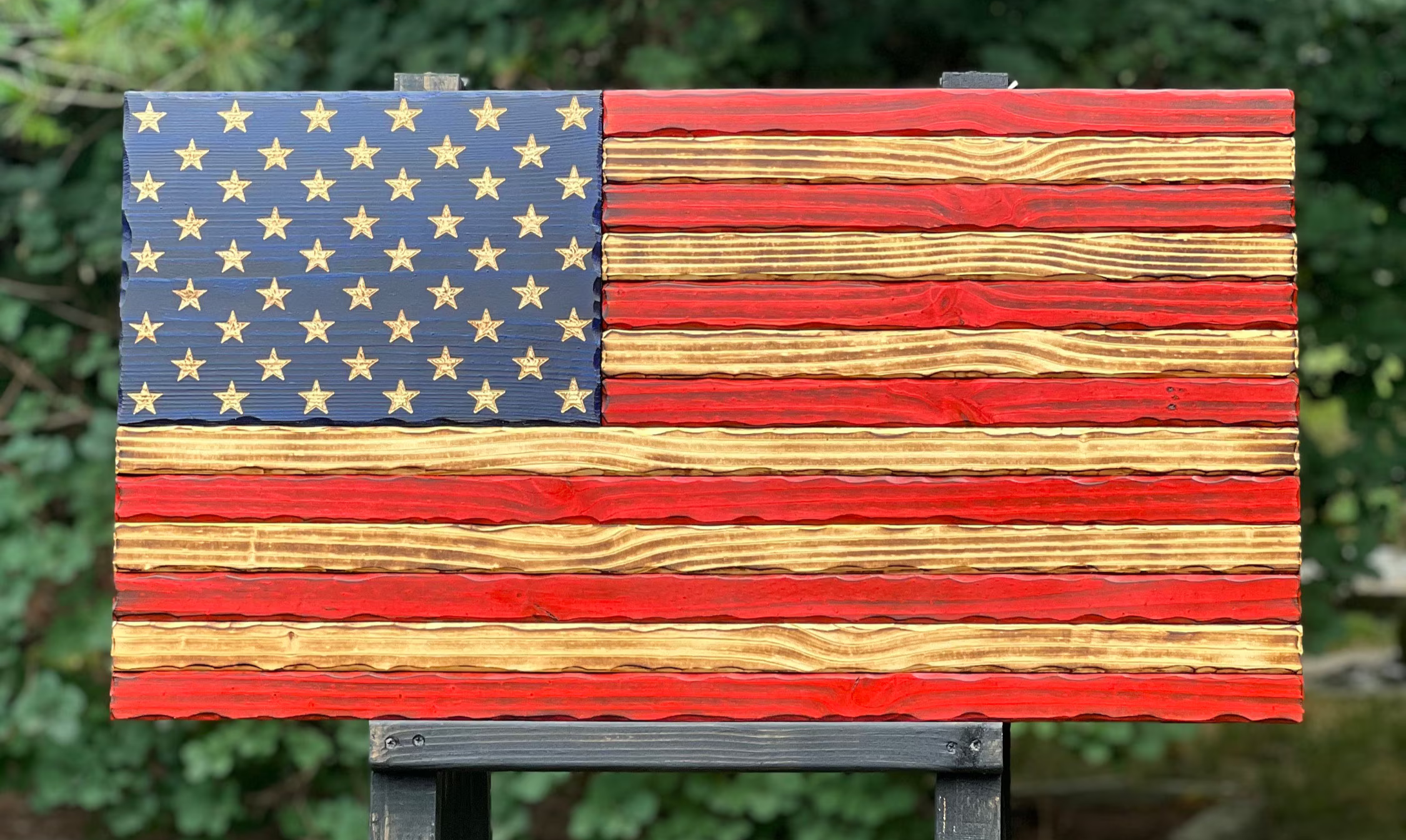 Wooden American flag on a stand with greenery in the background